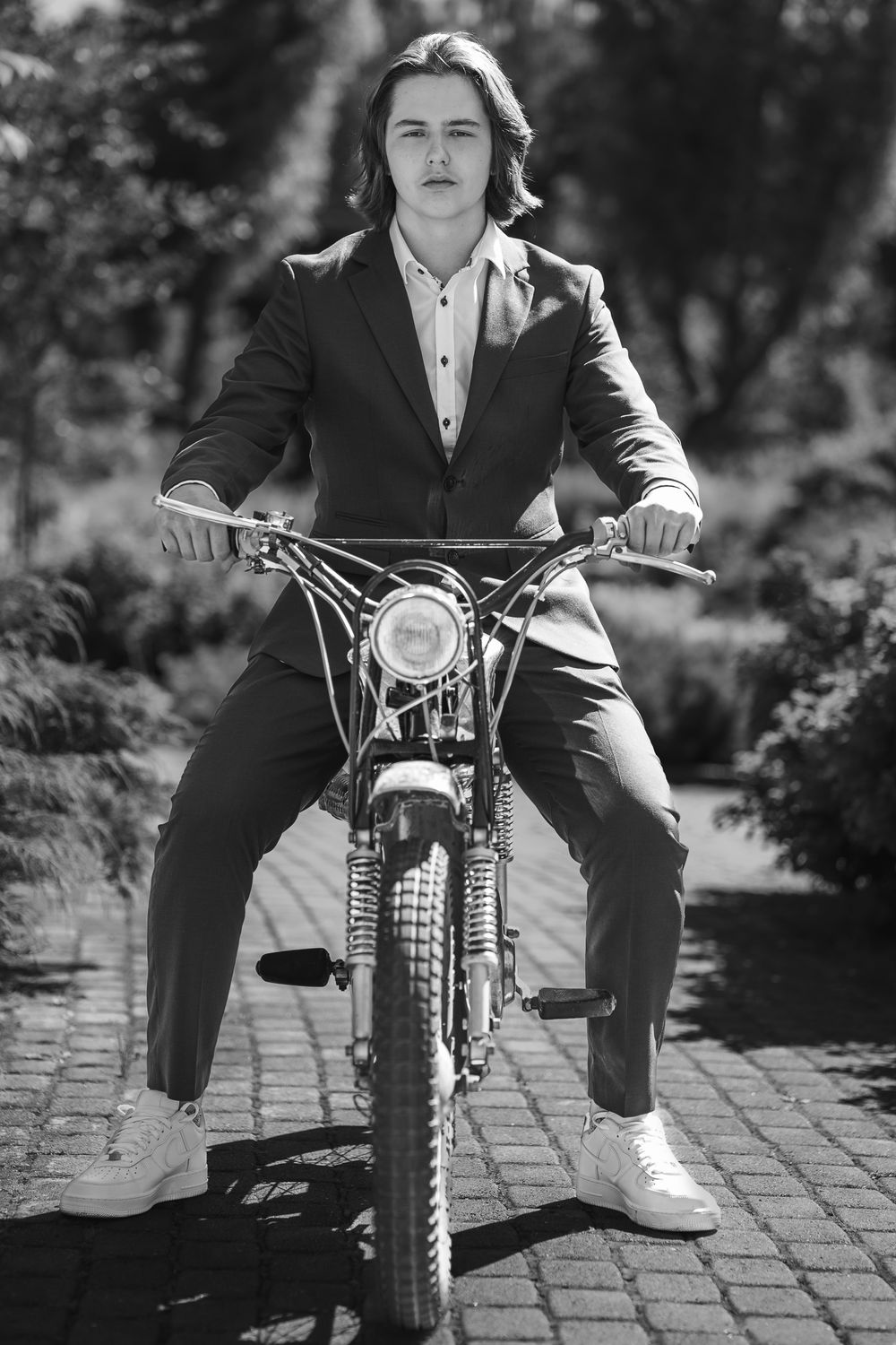 Black and white portrait of a person in a suit sitting on a vintage motorcycle on a brick paved surface outdoors.