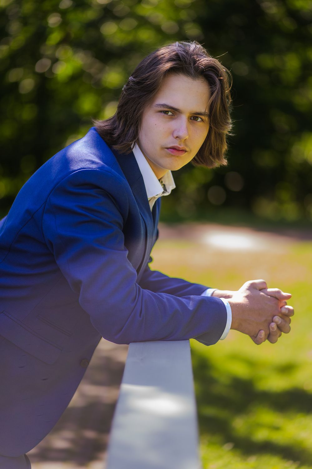 Portrait of someone in a stylish blue suit leaning against a railing in a sunny outdoor setting with bokeh background.