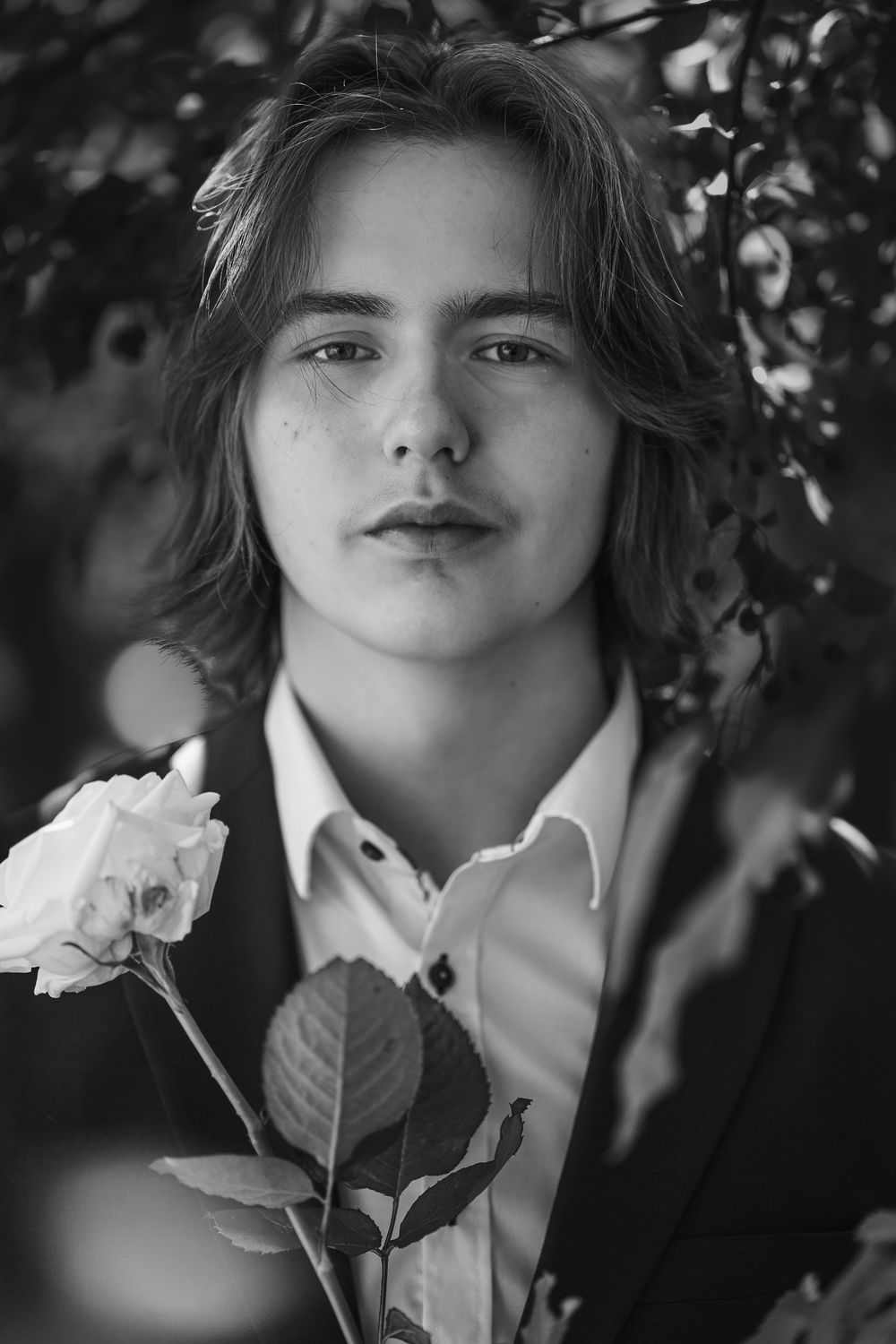 Black and white portrait of young person holding white rose against leafy background.