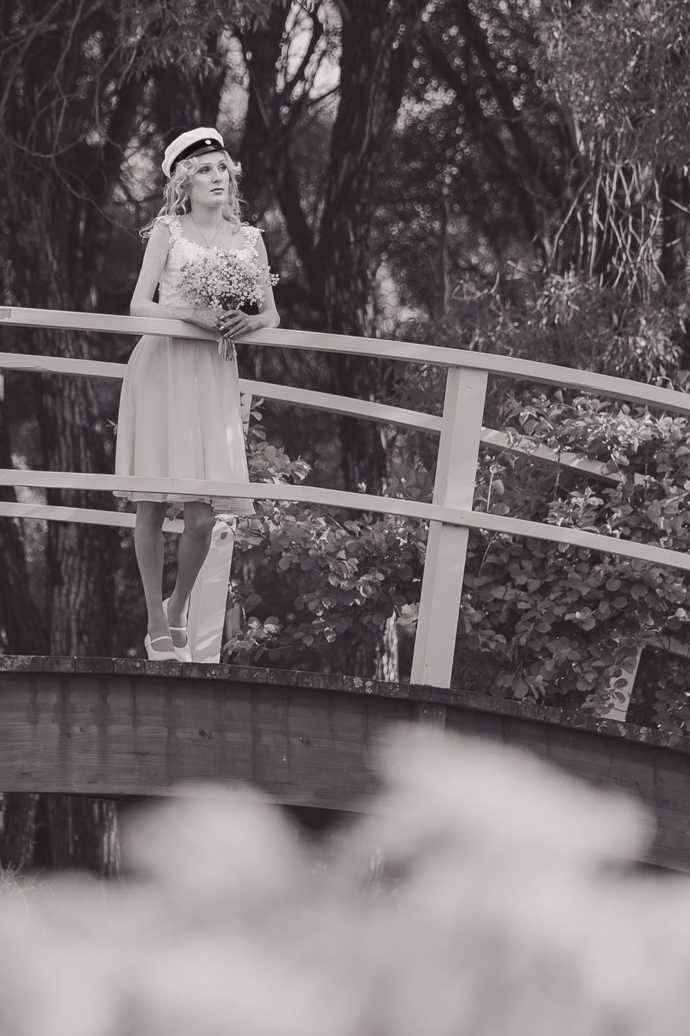 Black and white photo of someone in a white dress standing on a bridge in a garden setting.