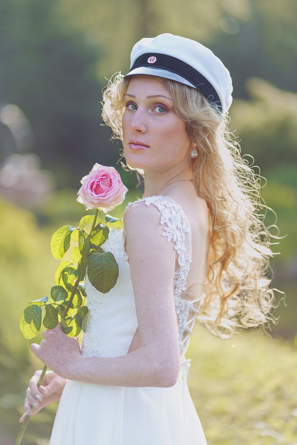 Person in white lace dress and cap holds pink rose in sunny garden setting at sunset.
