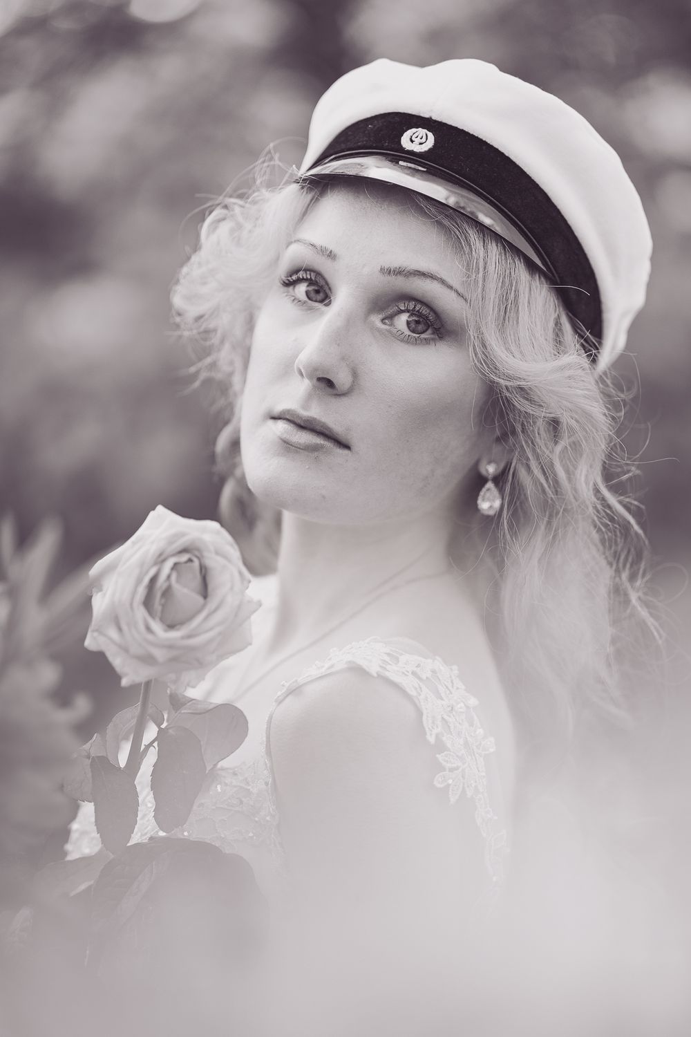 Romantic black and white portrait shows a woman in a white dress and nautical cap holding a rose.