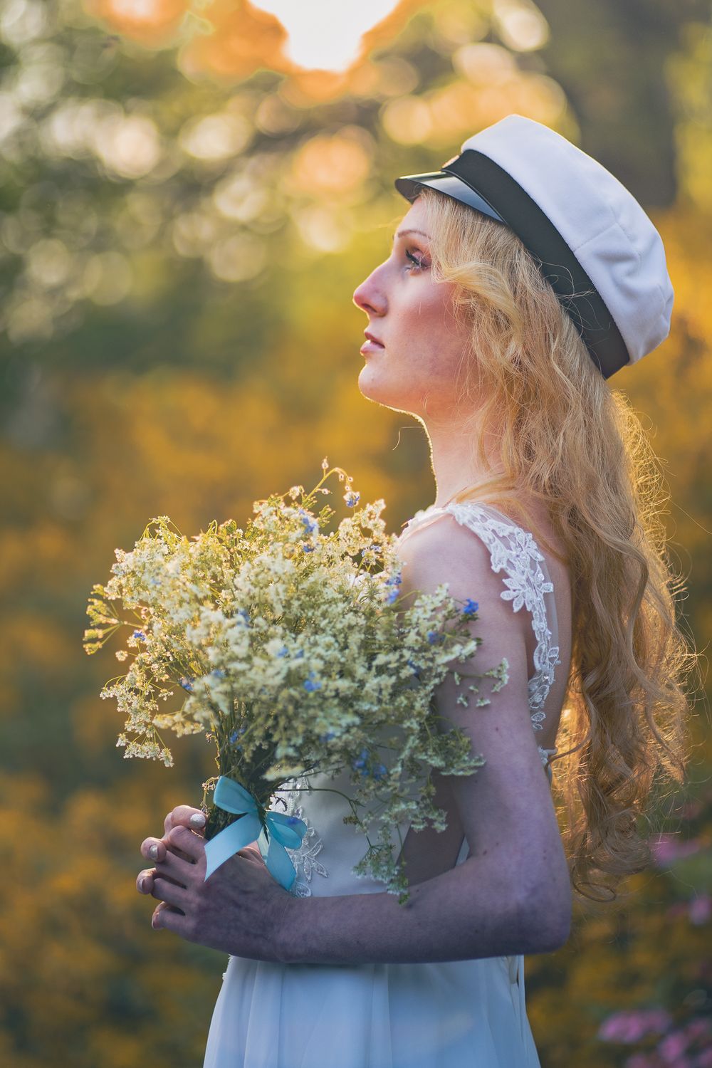 Dreamy profile portrait of someone in white dress and hat holding wildflowers against golden autumn bokeh background.