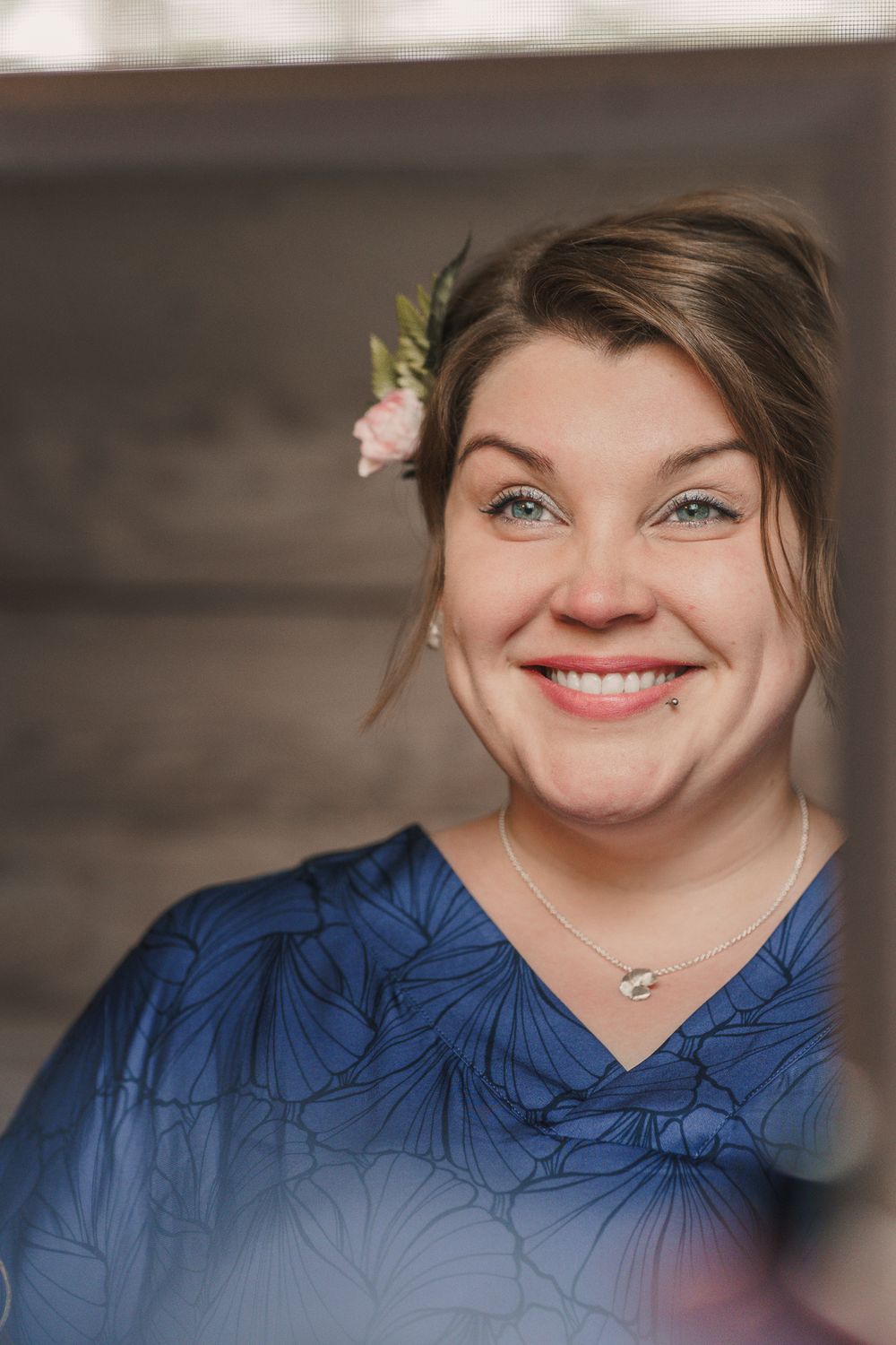 A joyful smile brightens the portrait as a delicate pink flower adorns styled hair, complemented by a blue patterned top.