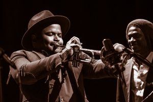 Trumpet player performing with fellow band mate looks on smiling, in black and white.
