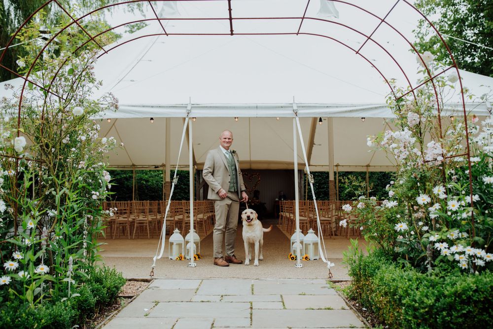 Father with dogs walks under white tent archway decorated with climbing flowers. Houchins wedding venue