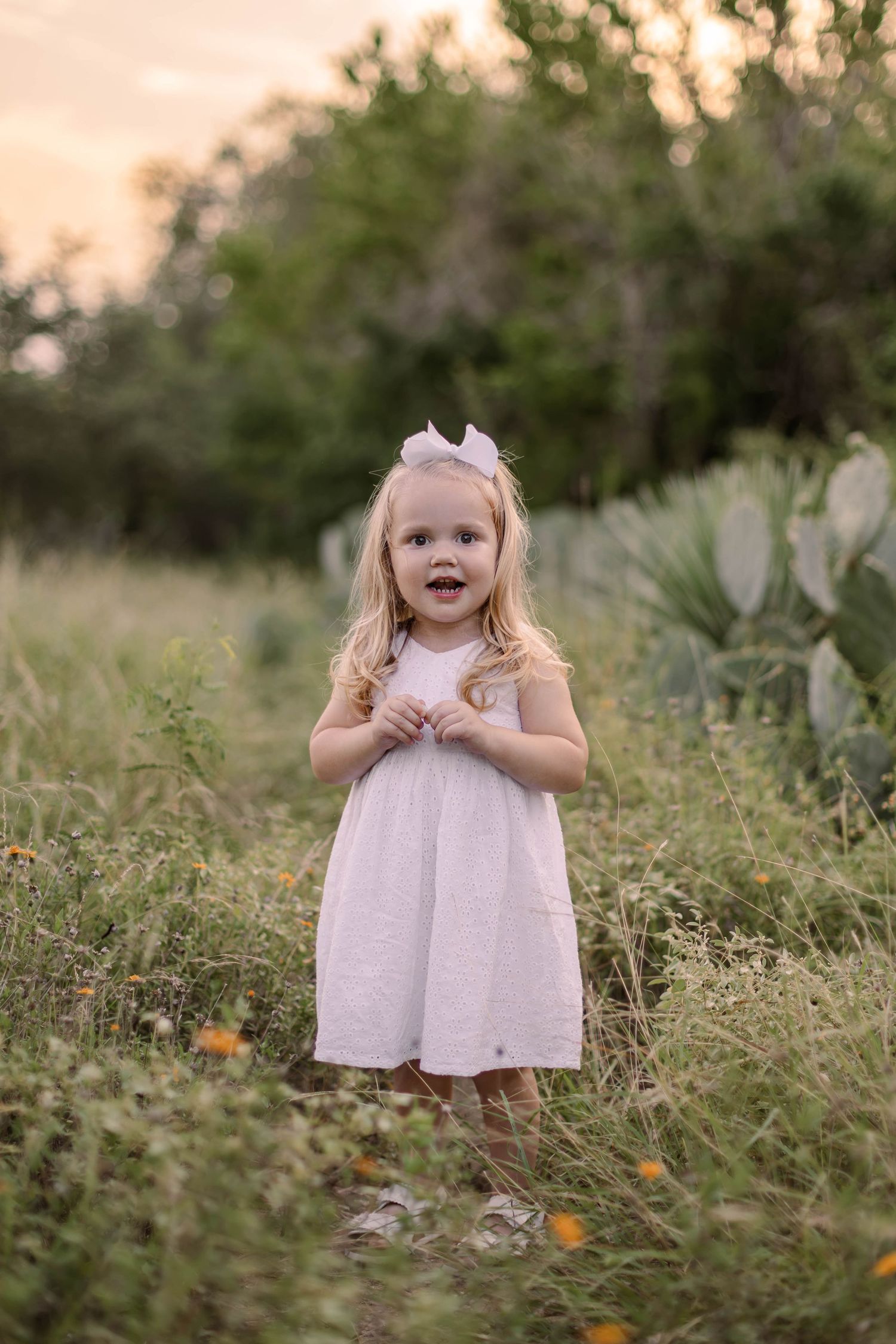 A young child in a peach-colored dress poses on grassy ground during sunset creating a warm golden atmosphere.