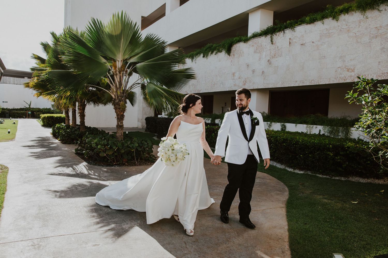 A tropical wedding photo with palm trees and white wedding attire along a garden path.