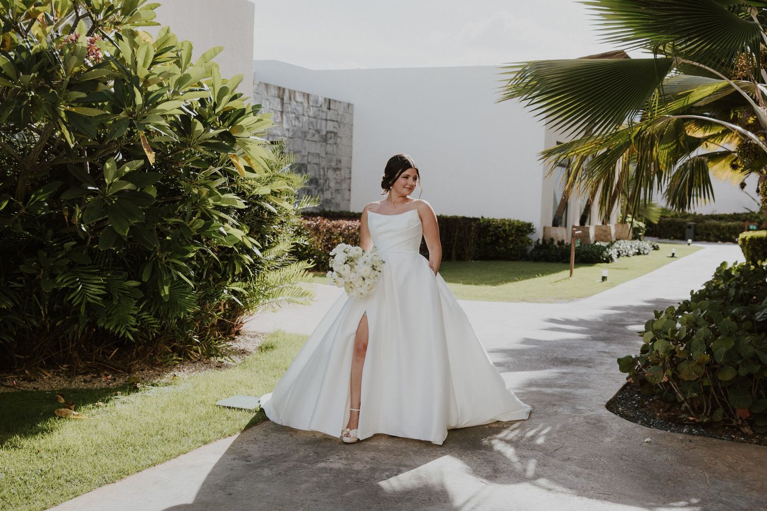 A white bridal gown with side slit flows elegantly along a palm-lined path on a sunny day.