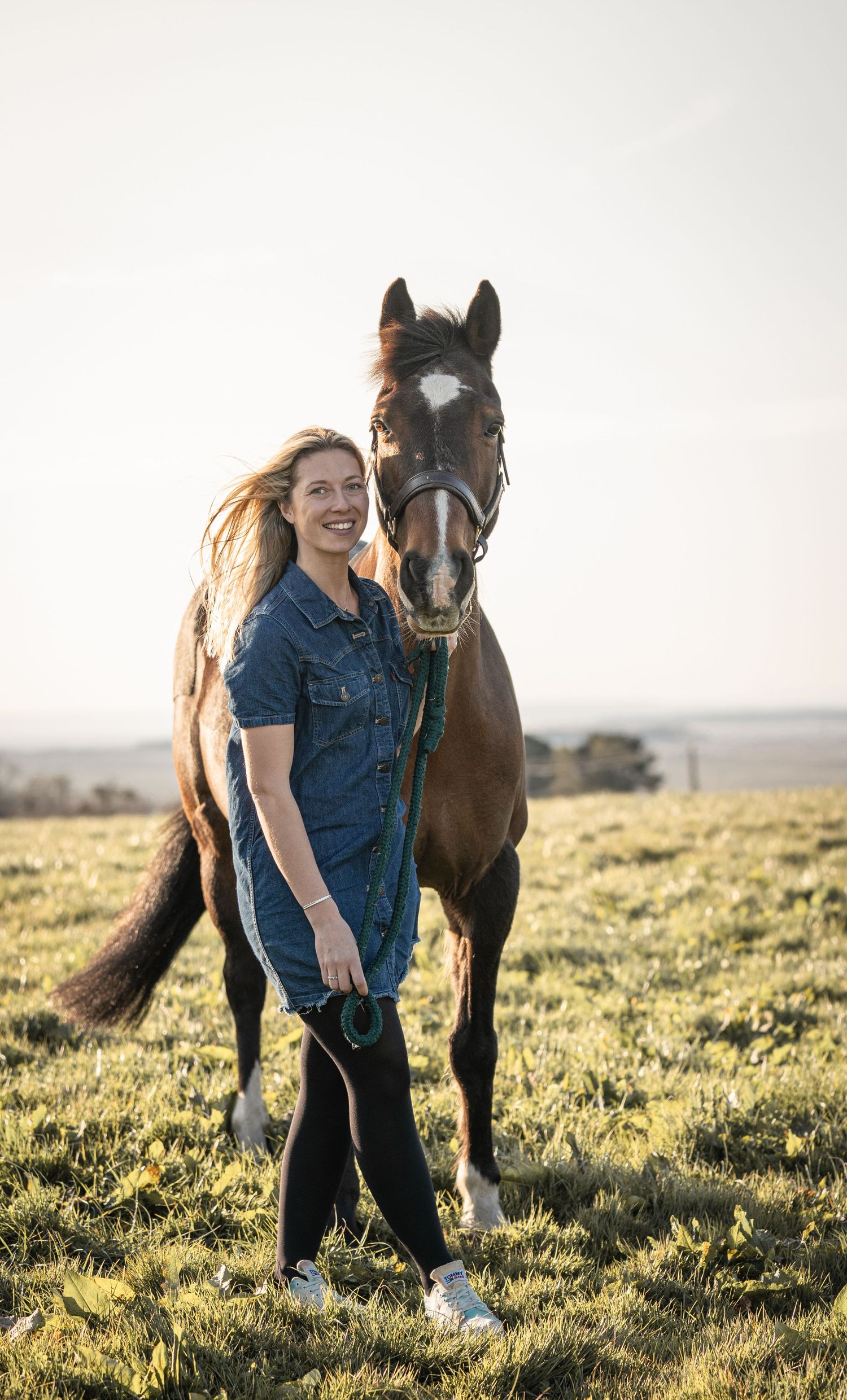 A rider on horseback moves through a sunlit field at dawn with houses visible in the background.