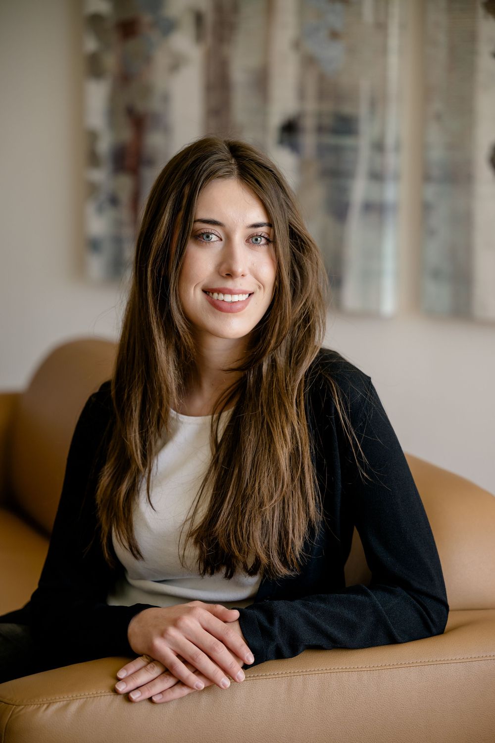Professional portrait of a smiling person with long brown hair wearing a black cardigan and white top, seated on a tan couch.