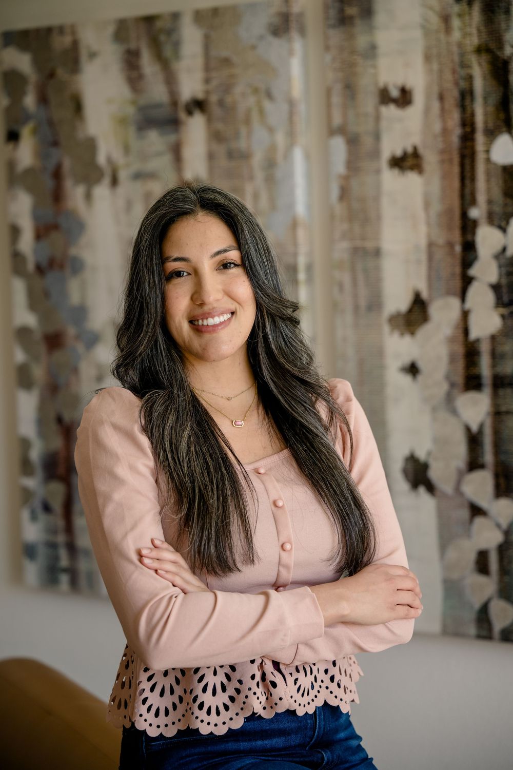 Indoor portrait of a woman in a pink lace-trimmed top and jeans posed in front of a textured wall.