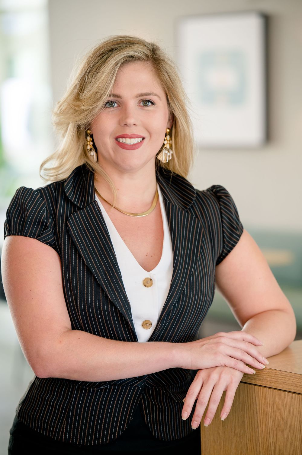 Professional business portrait of someone wearing a pinstriped blazer and white top with gold jewelry against blurred office background.