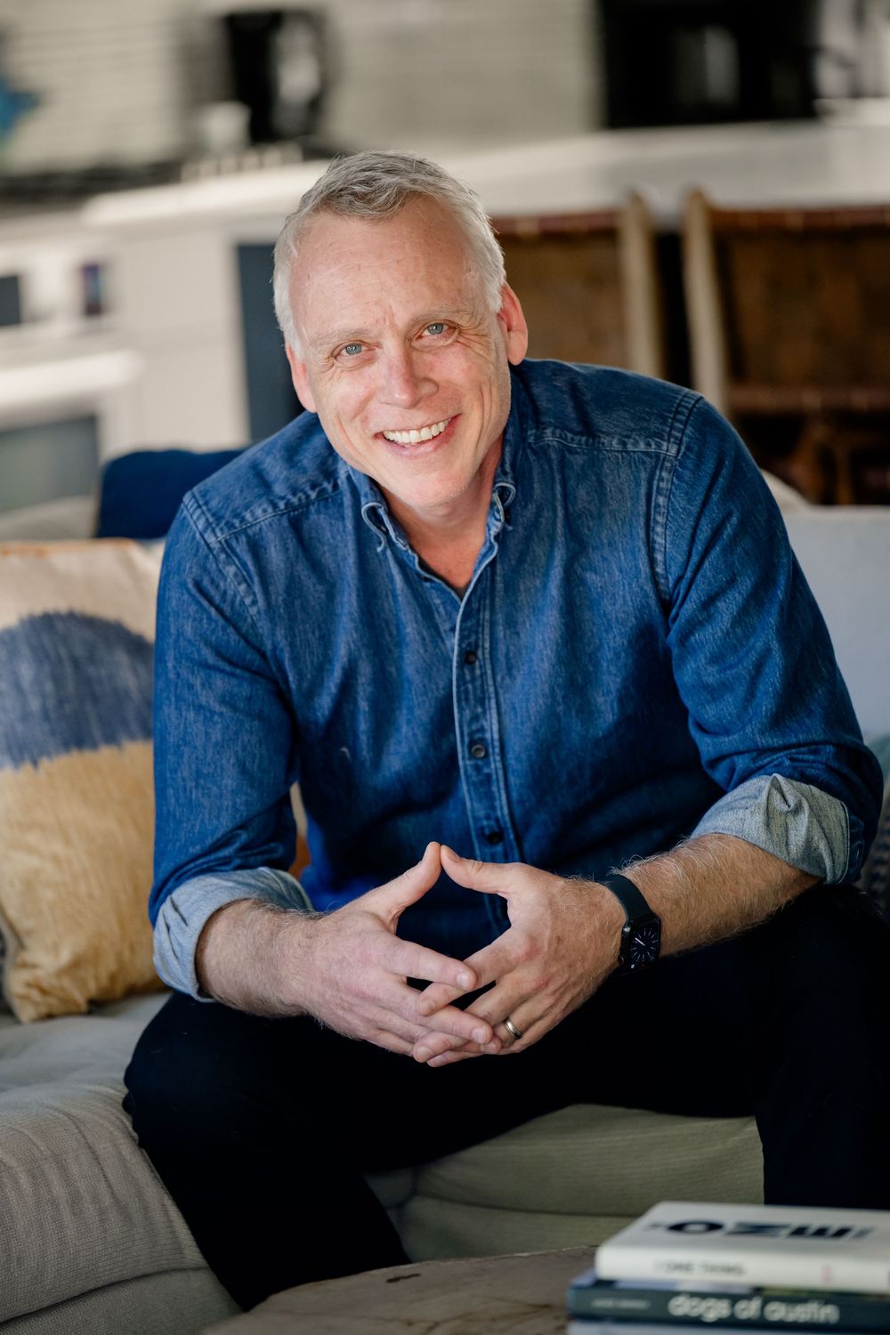 Person in denim button-down shirt sits casually on gray couch with warm smile in home setting.