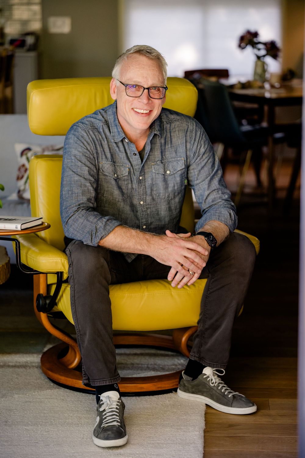A person wearing glasses and a gray button-down shirt sits smiling in a bright yellow leather armchair.