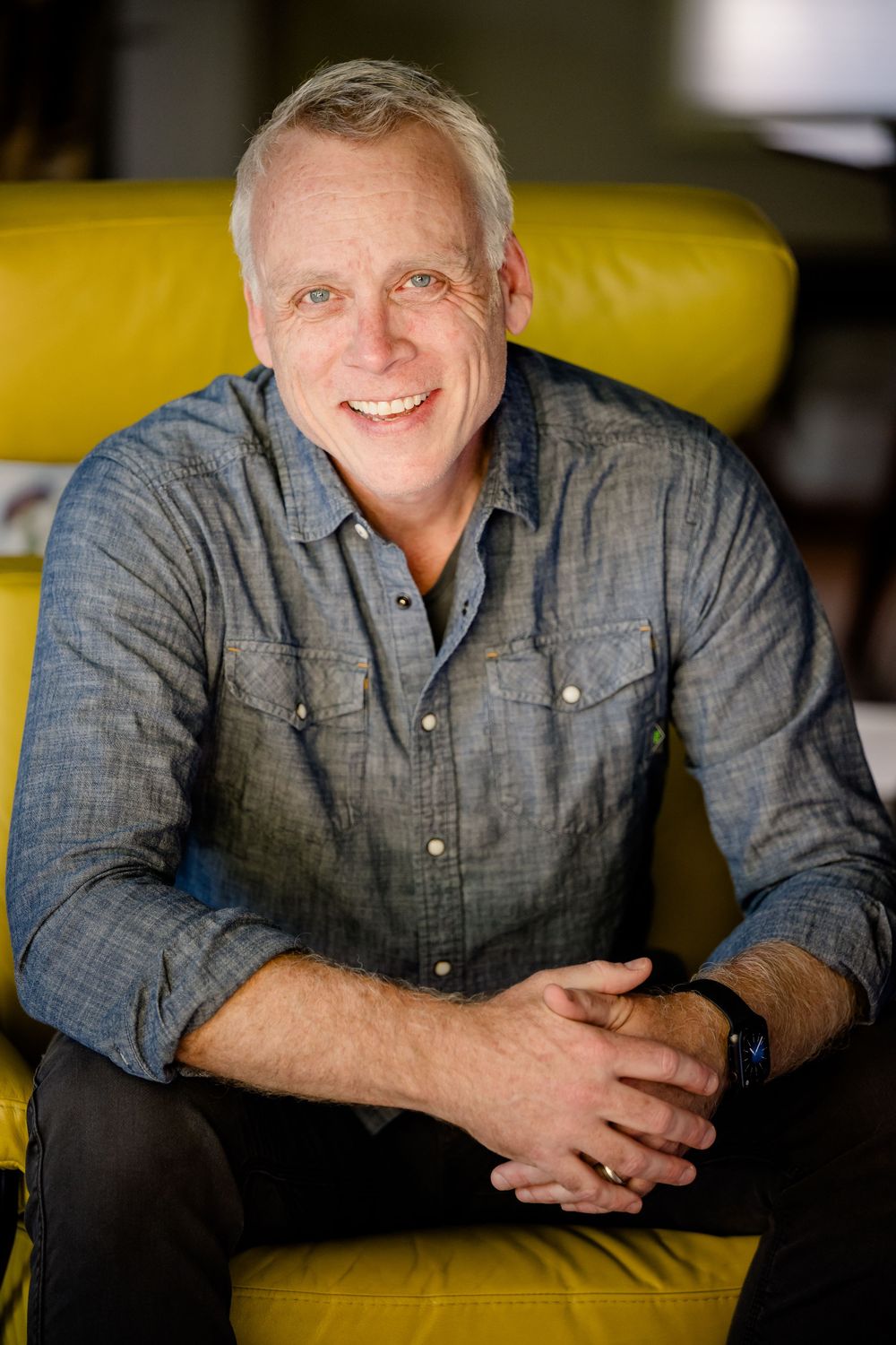 Professional headshot portrait of someone in a chambray button-down shirt against yellow background with warm lighting.