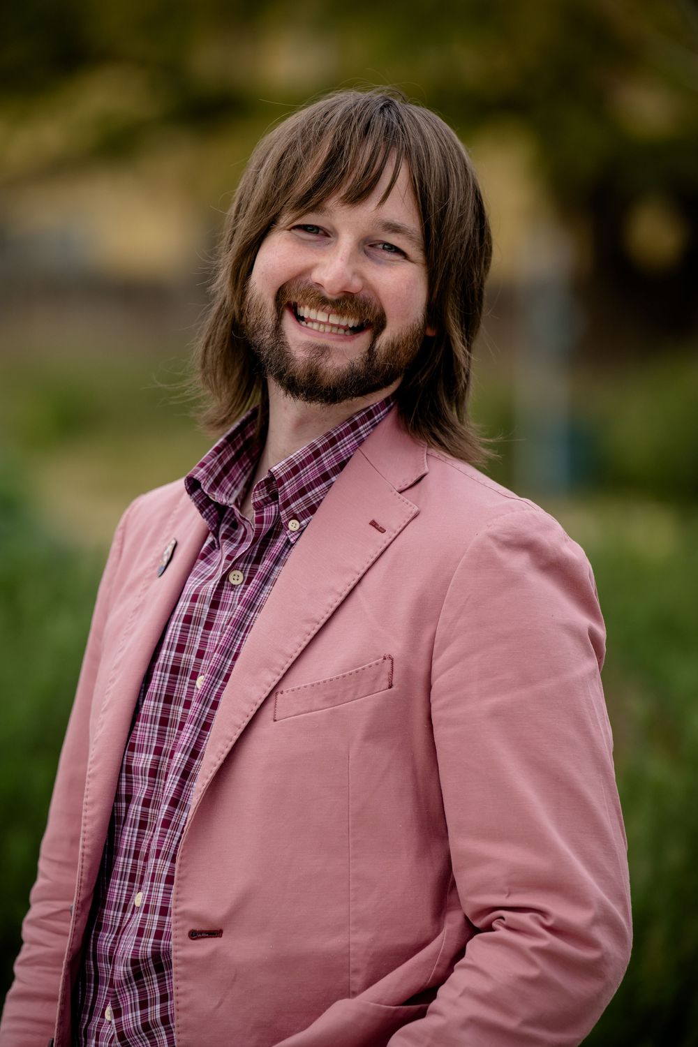 A professional portrait shows someone wearing a pink blazer and plaid shirt with long hair and a beard.