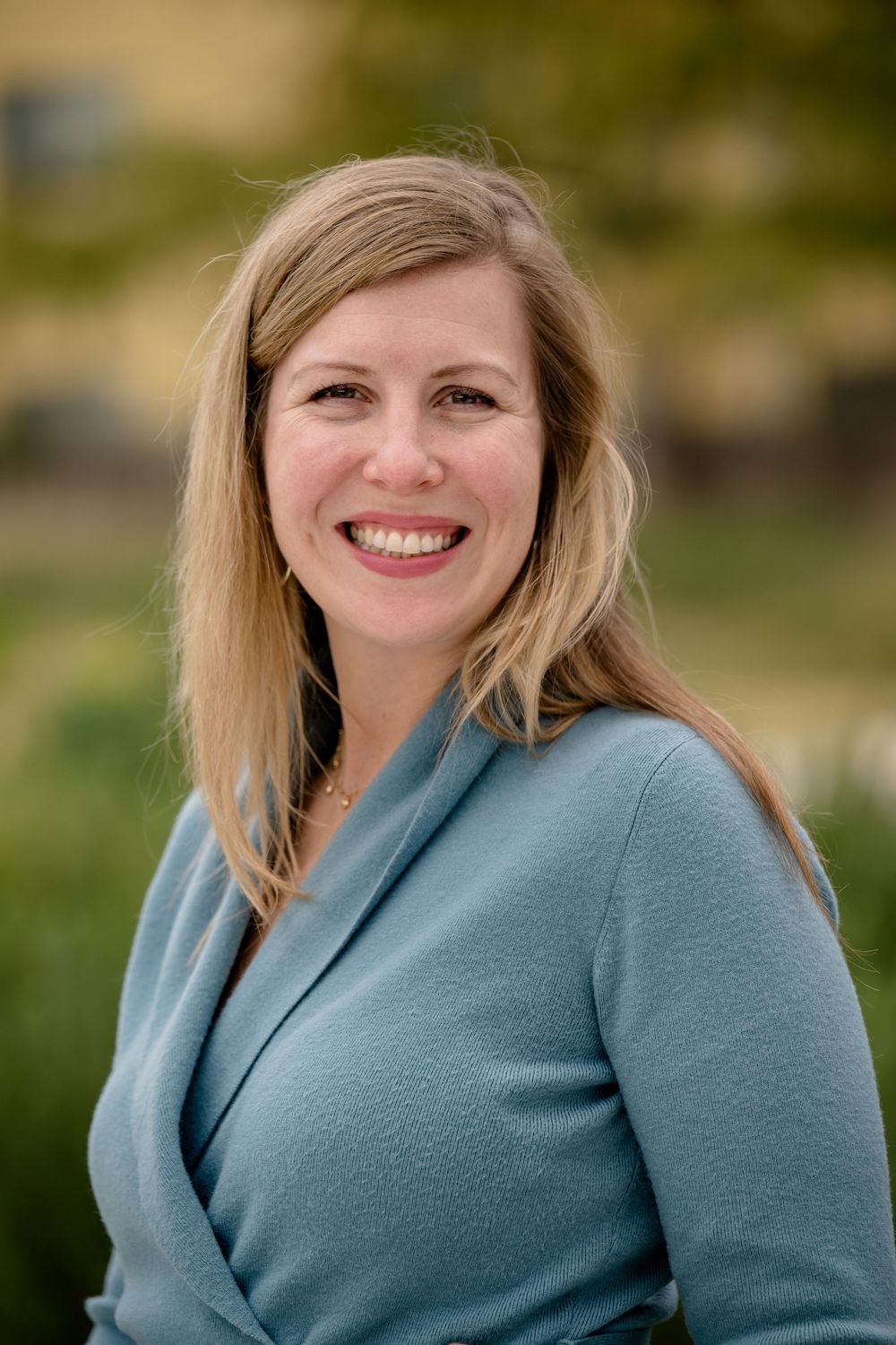 Professional headshot of a smiling woman in a teal sweater outdoors with a blurred natural background.