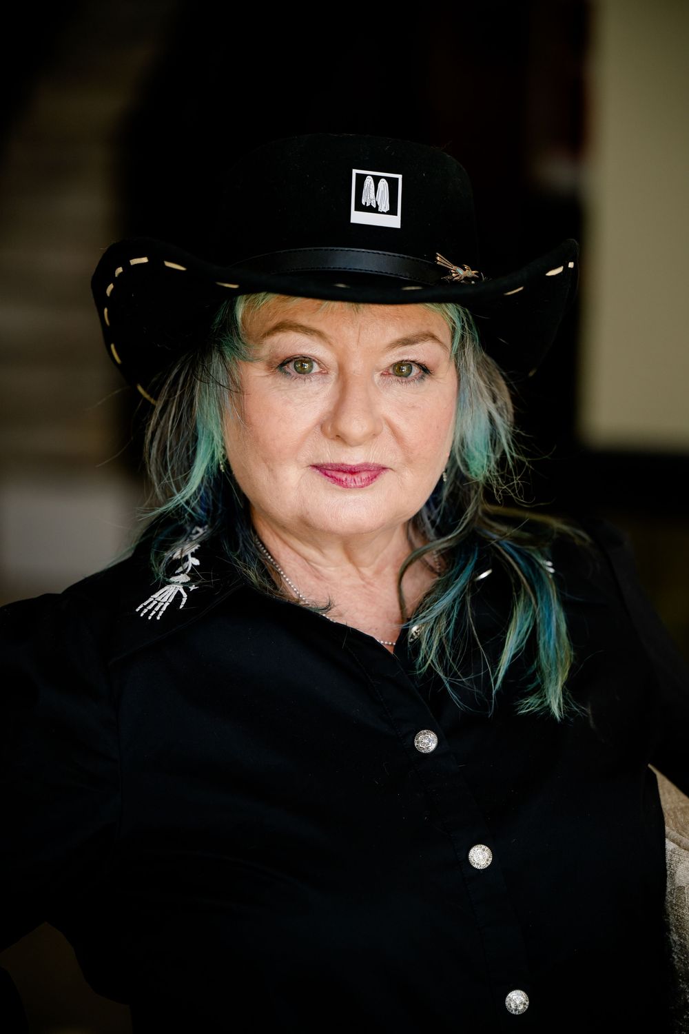 Person wearing black cowboy hat with blue-tinted hair and black button-up shirt smiles warmly at camera.