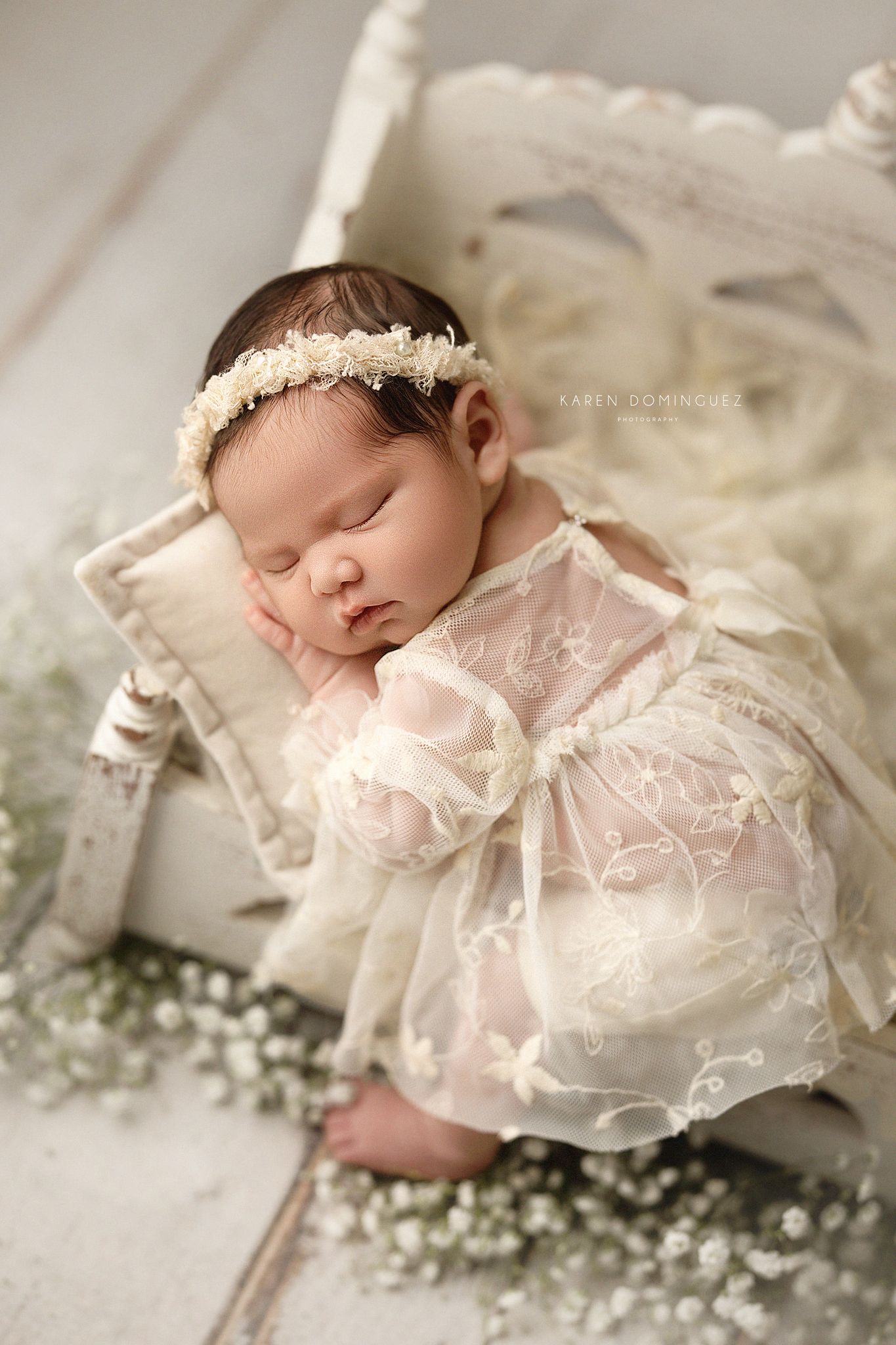 Sleeping newborn dressed in delicate white lace dress and floral headband lying on soft fabric with baby's breath flowers.