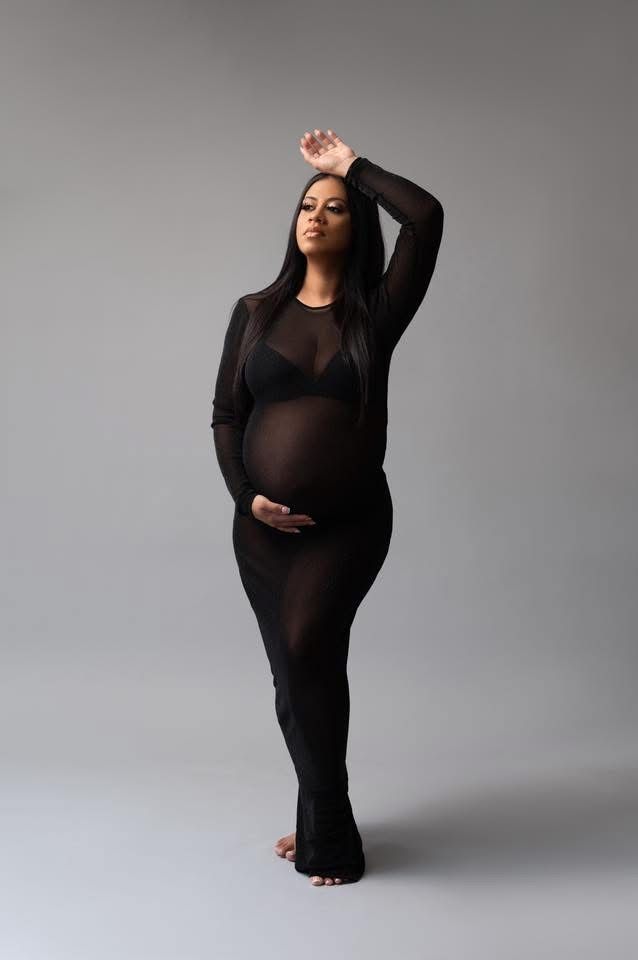 Pregnant woman in sheer black bodysuit poses gracefully against gray backdrop in artistic maternity photo.