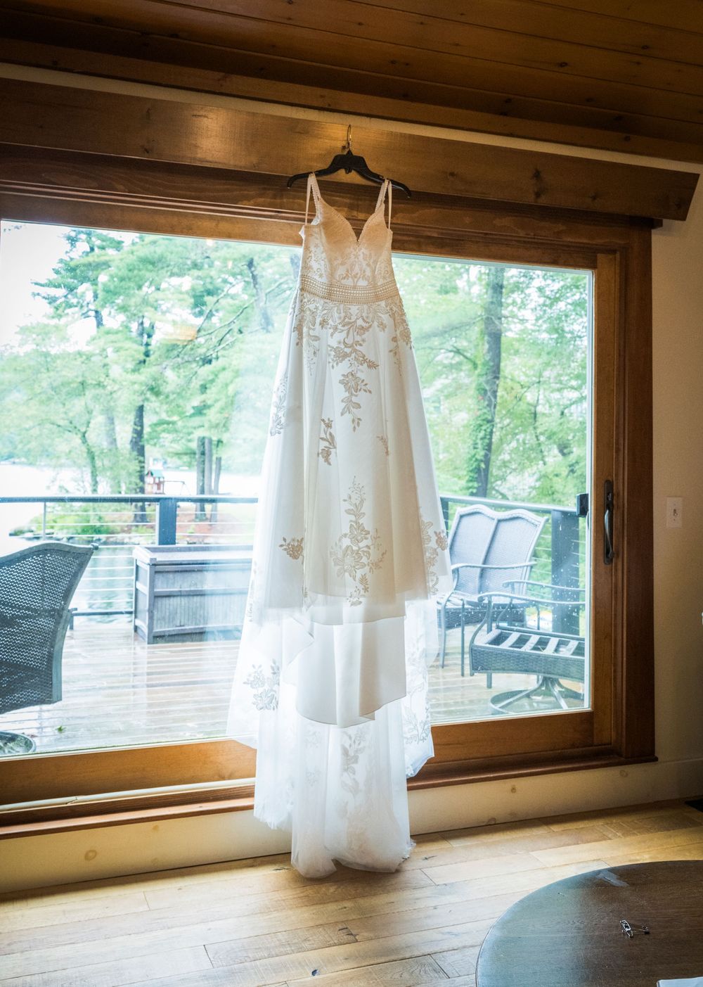 White wedding dresses hanging from wooden beams in a rustic room with large windows overlooking green trees outside.
