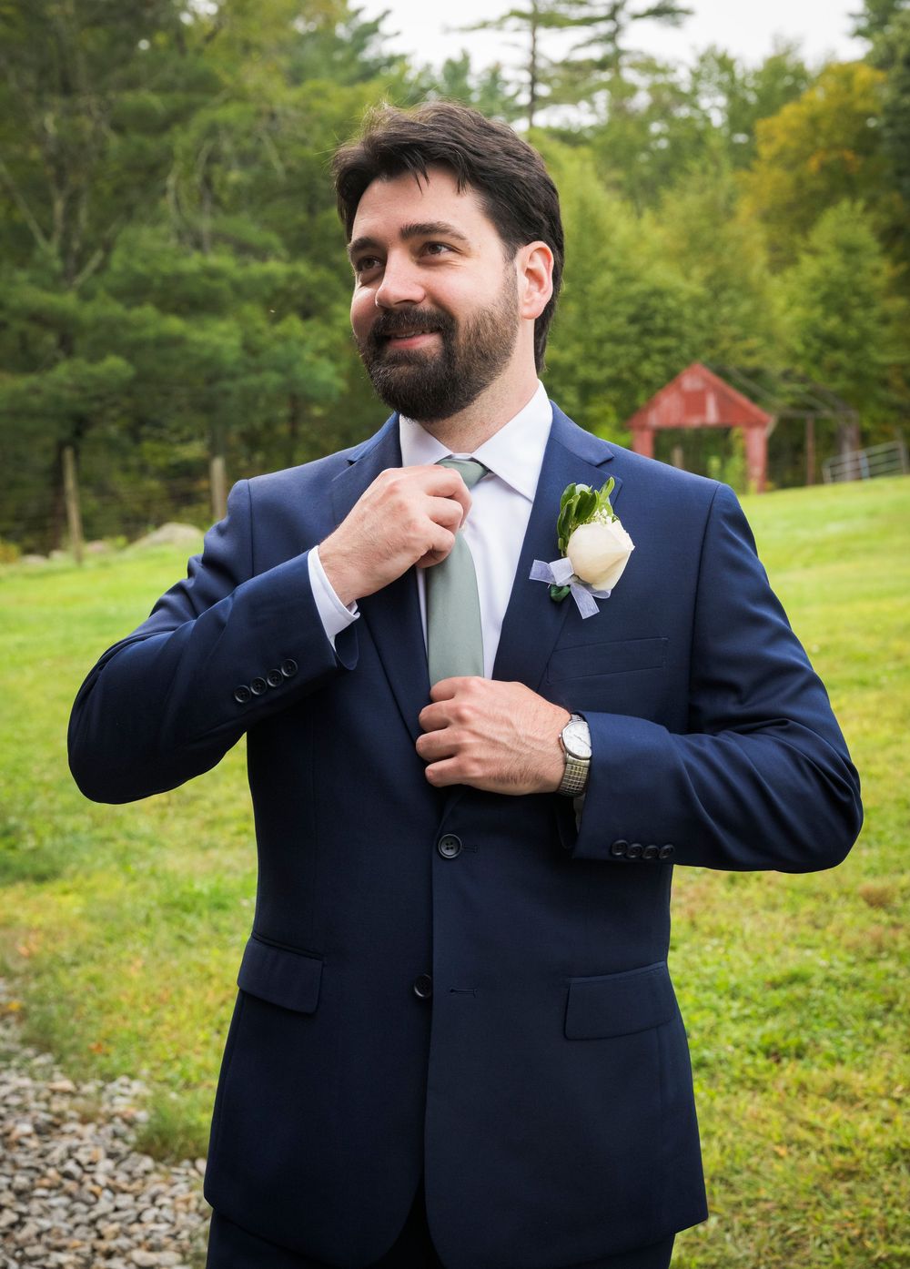 Groom in navy suit adjusts his tie while getting ready outdoors with green foliage in the background.