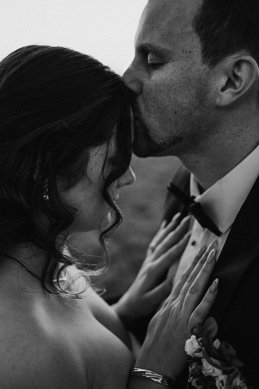 An intimate black and white wedding photo showing a tender forehead kiss between newlyweds in formal attire.