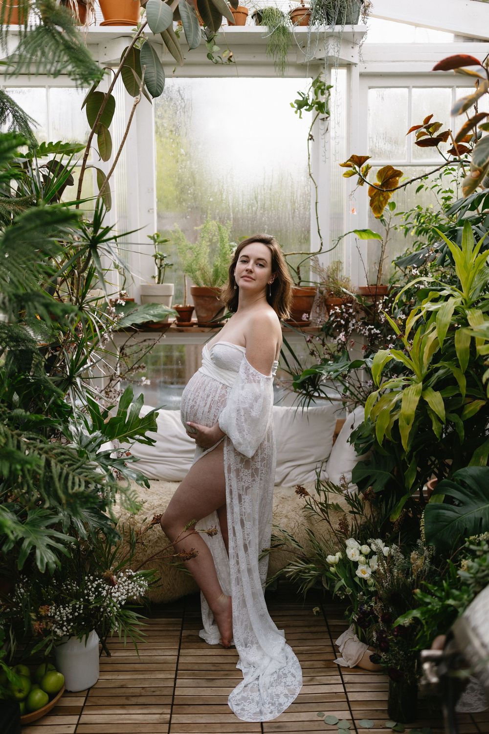 Dreamy boudoir photoshoot featuring white lace dress surrounded by lush indoor plants and natural light in greenhouse setting.