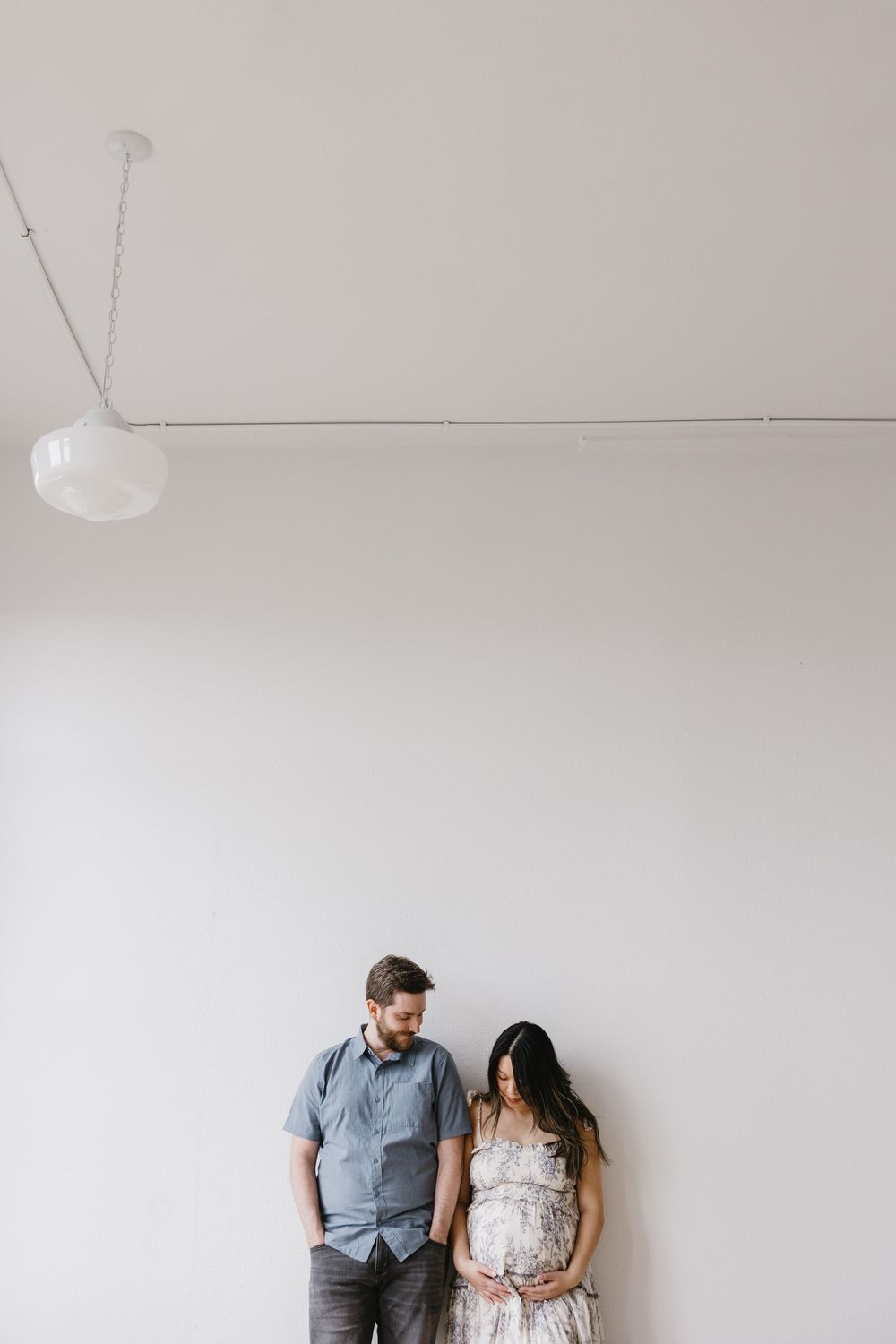Simple minimalist maternity photo against white wall with hanging pendant light fixture.