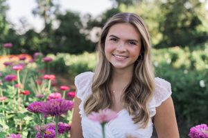 A woman wearing a white top smiles while posing among pink flowers and greenery in a garden setting.