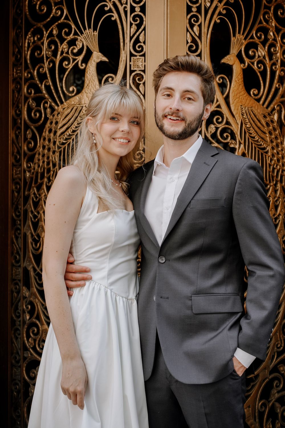 A couple poses in formal wedding attire against an ornate gold art deco door with intricate peacock designs.