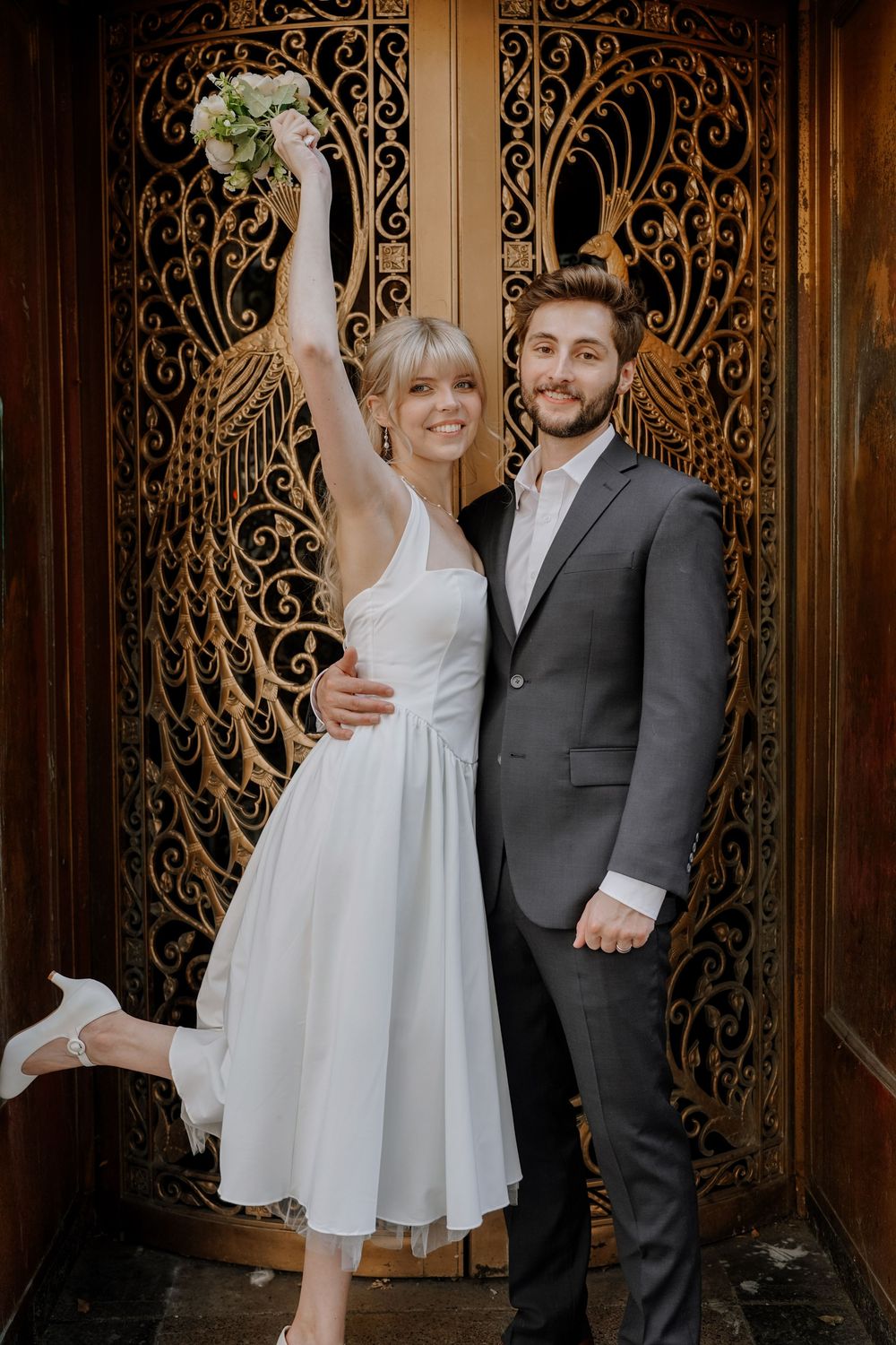 Couple poses in front of ornate wrought iron door, with person in white dress playfully lifting one leg.