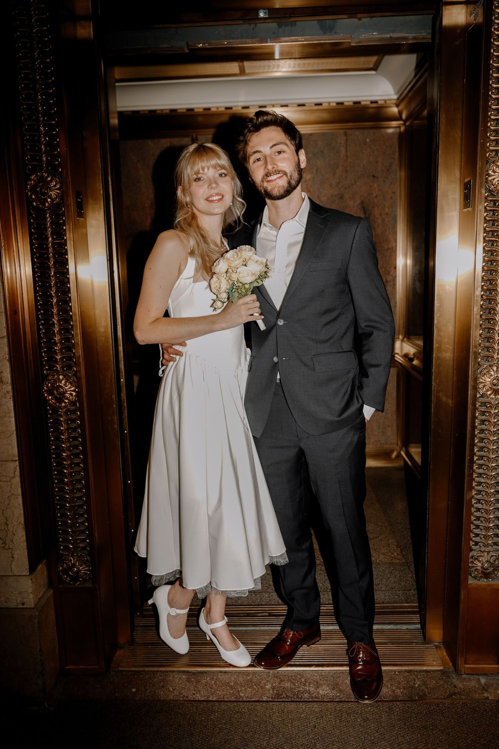 A romantic wedding couple poses in an ornate hallway with dark wood panels and vintage elevator details.