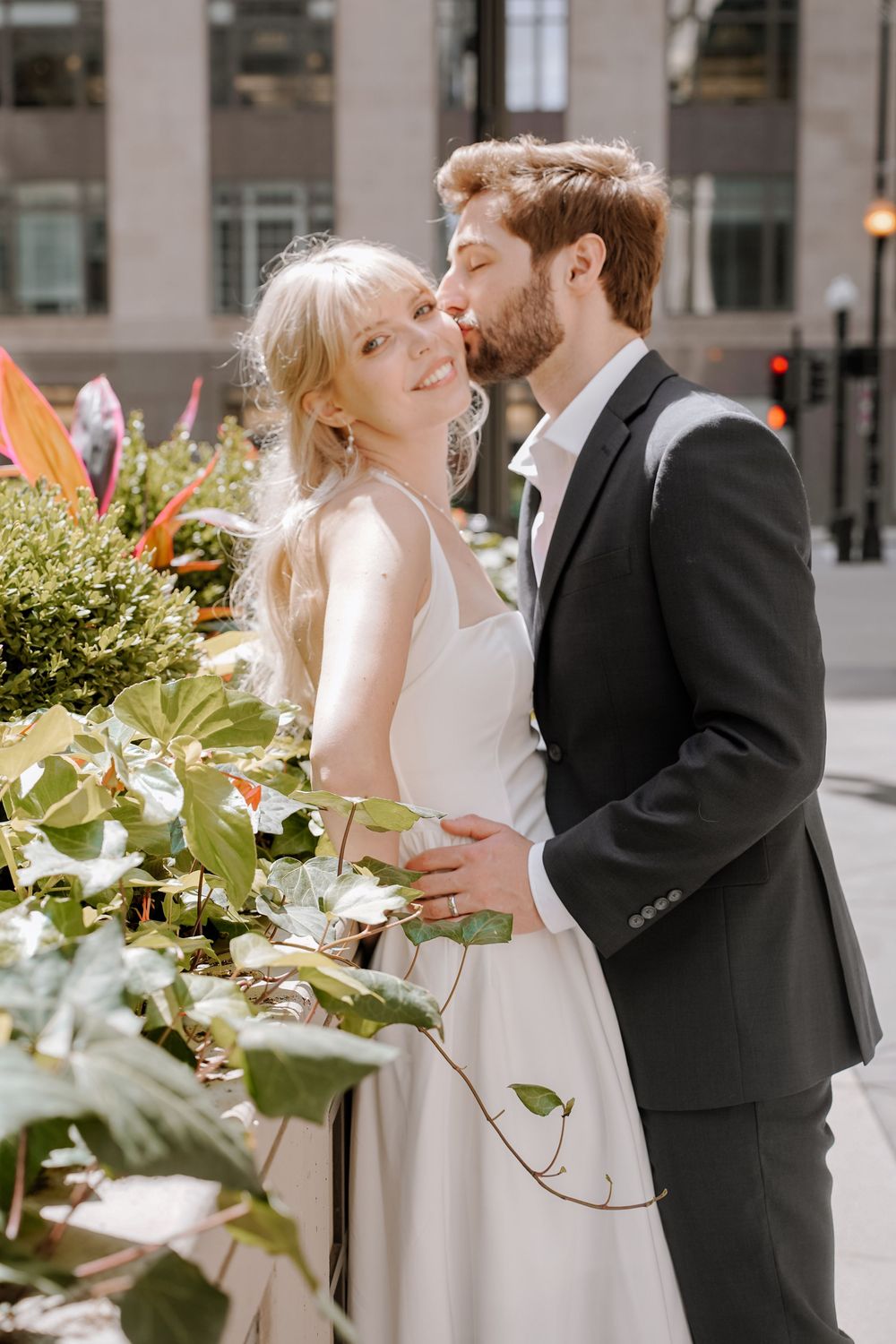 A romantic wedding couple embraces among decorative greenery and flowers in an urban setting.