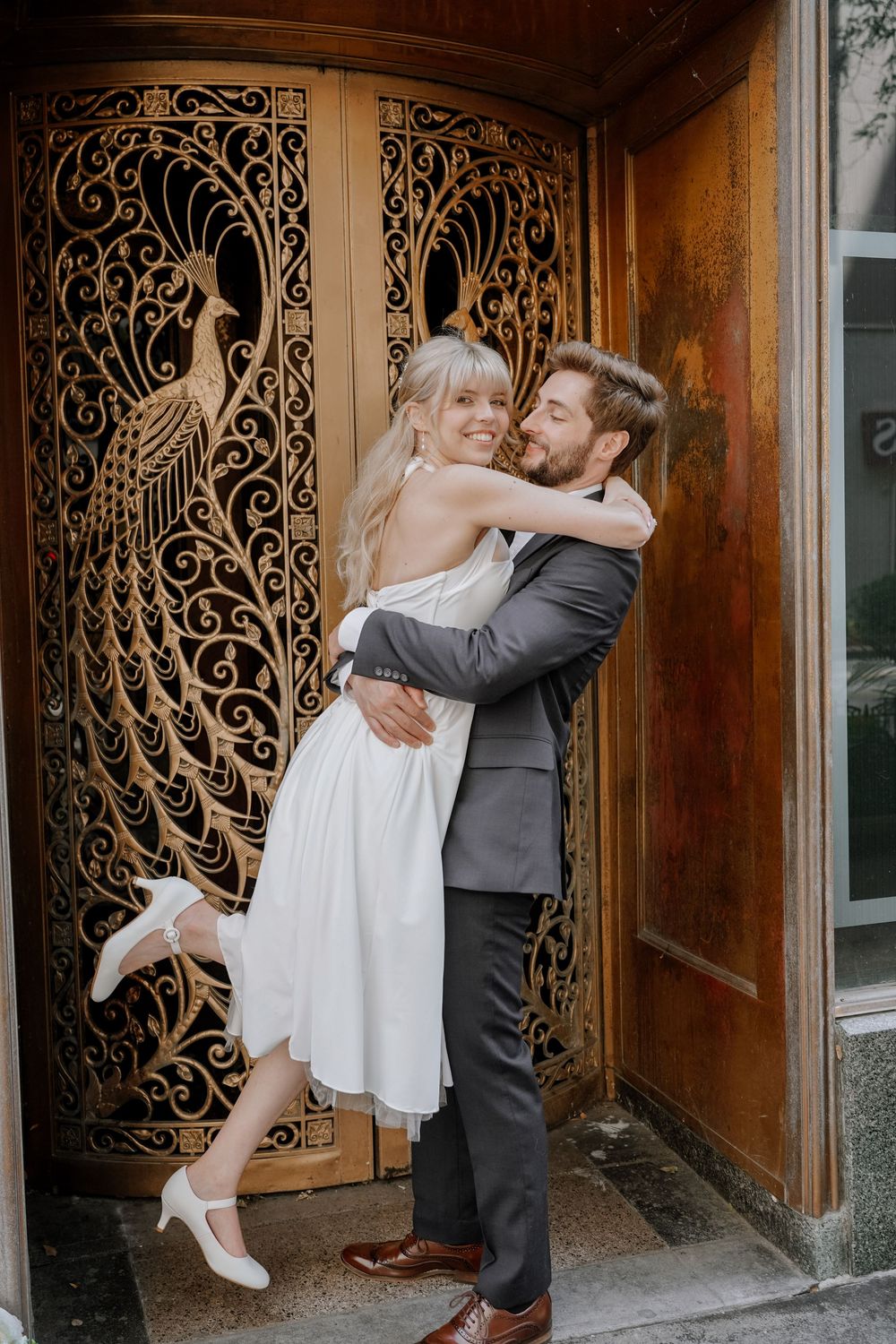 A newlywed couple shares an intimate moment in front of an ornate art nouveau door with peacock designs.