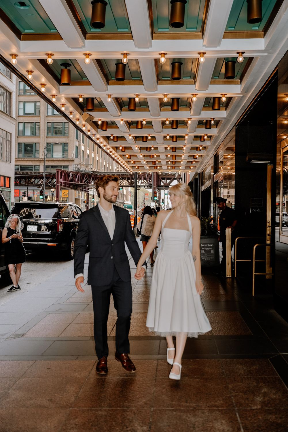 A couple dressed in formal attire walks together through an elegant hall with coffered ceilings and warm lighting.