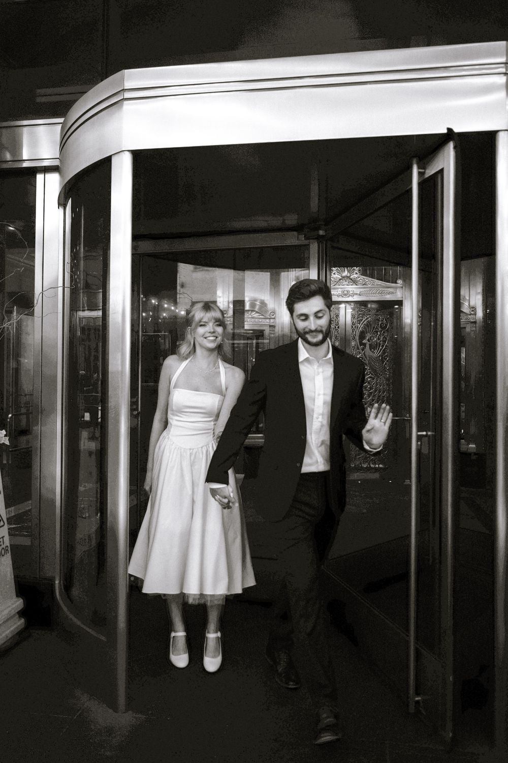 A couple in wedding attire stands in a revolving door entrance in black and white photography.