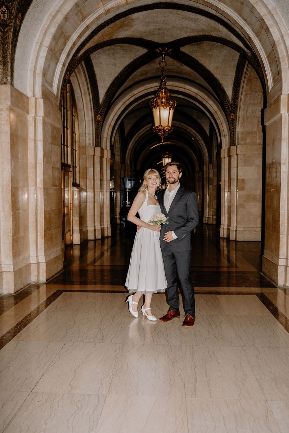 A formal couple stands together in a grand stone archway corridor with ornate architectural details.
