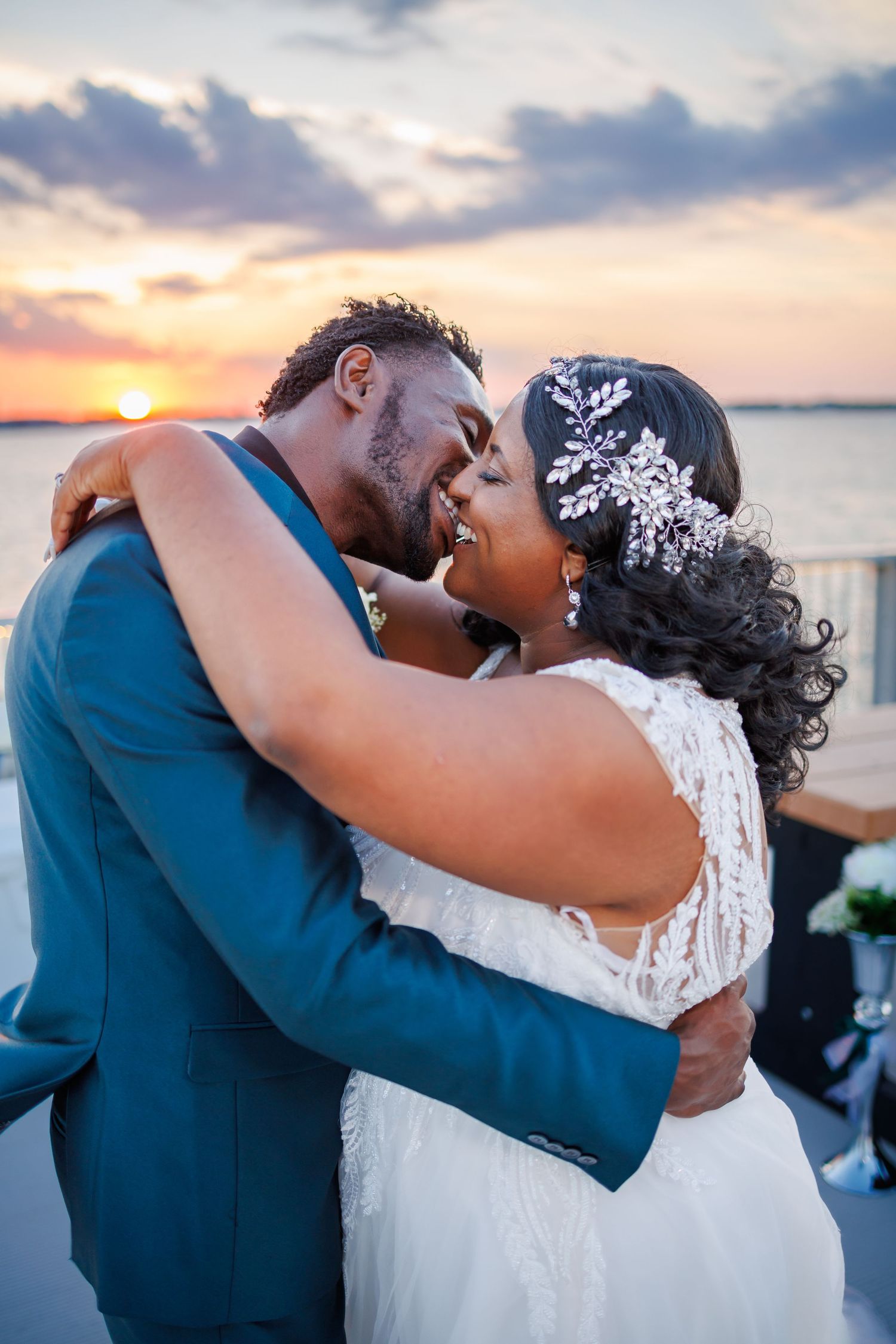 A couple in wedding attire share an intimate kiss at golden hour by the water's edge as the sun sets.
