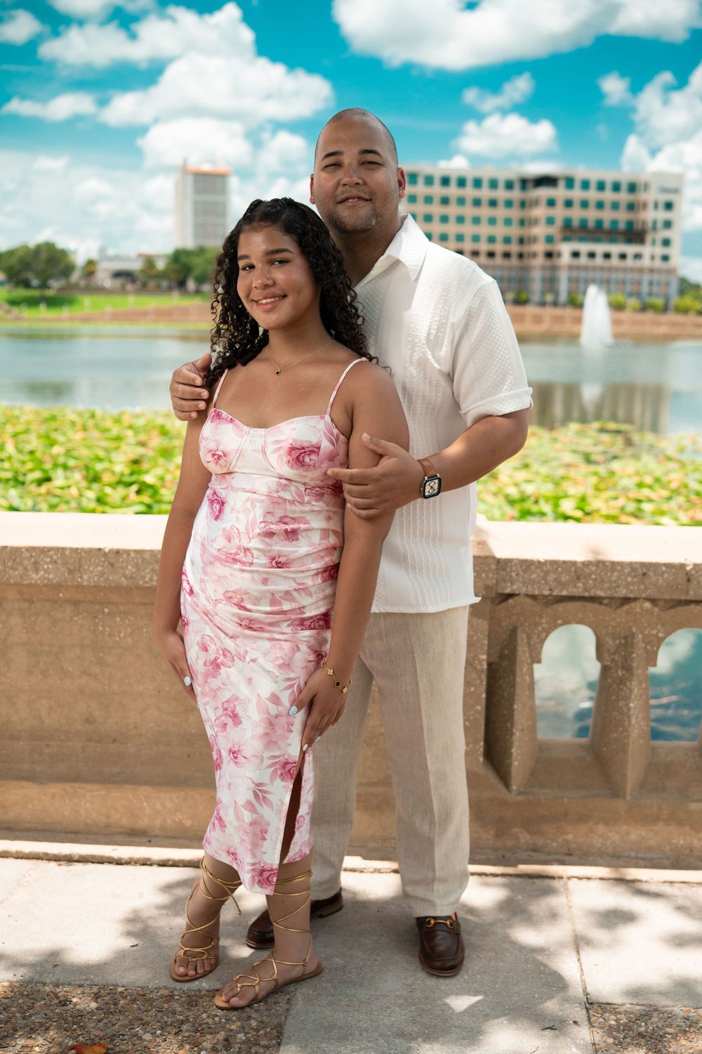 A couple poses together on a scenic balcony overlooking a lake with fountains and lily pads on a bright sunny day.