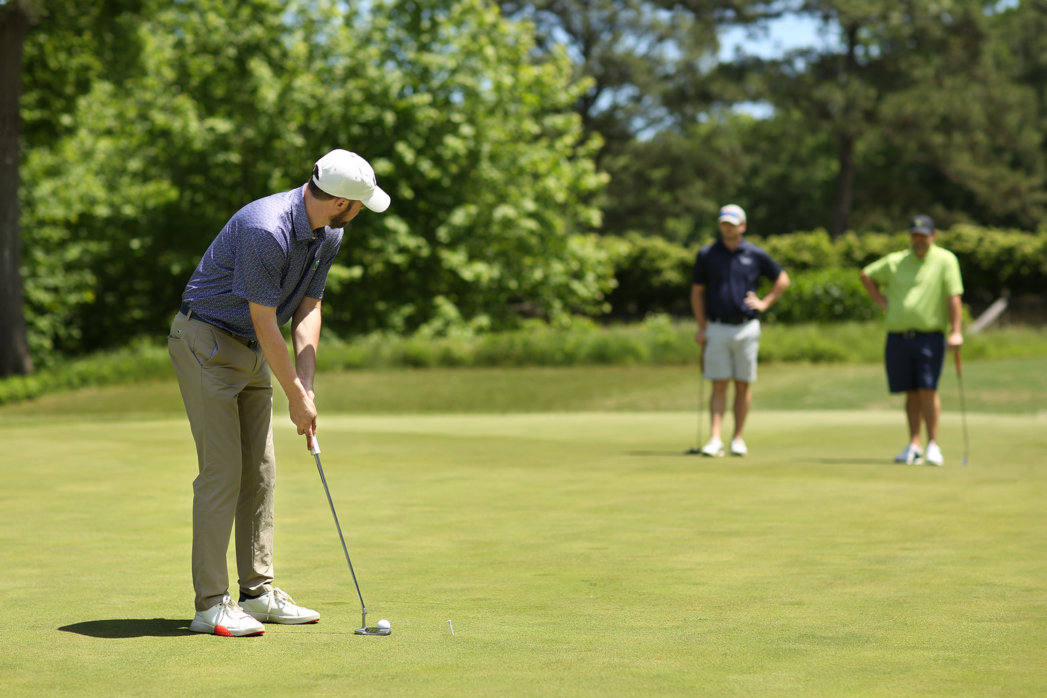 A golfer prepares to putt on a green putting surface while other players watch in the background.