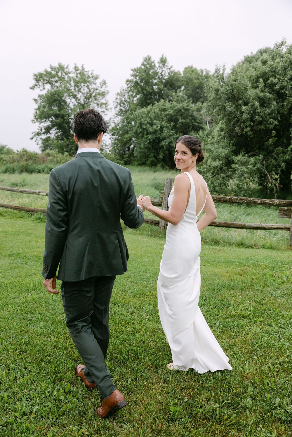 A couple in wedding attire walks together on a lush green lawn with trees in the background.