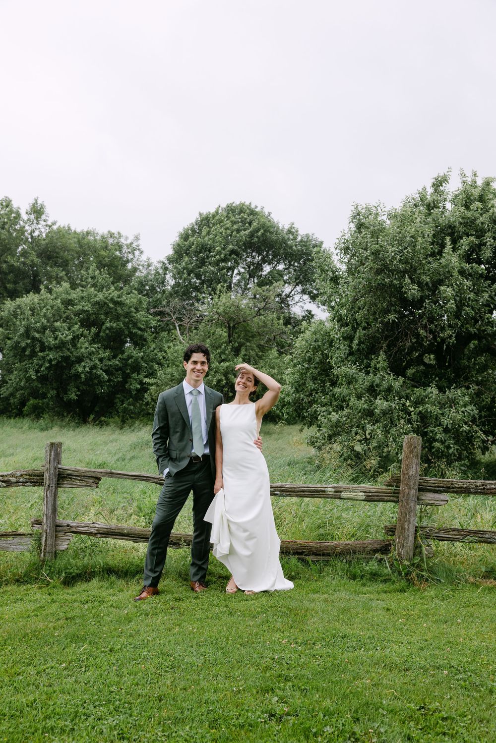 A couple in wedding attire poses playfully along a rustic wooden fence in a lush green field.