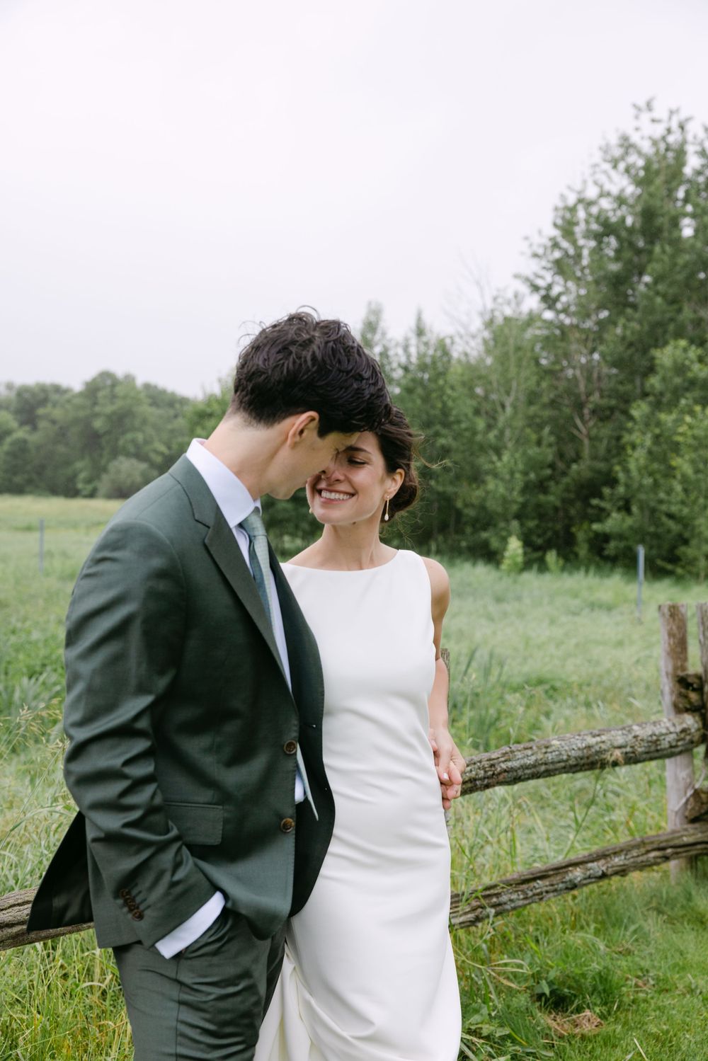 A bride and groom share an intimate moment by a rustic wooden fence in a pastoral setting.