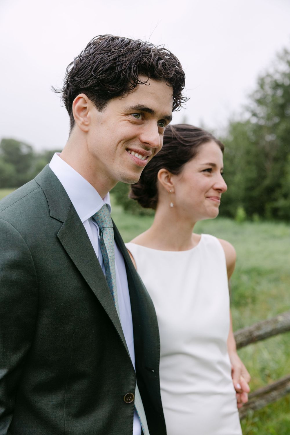A couple in wedding attire smiles while standing outdoors with greenery in the background.