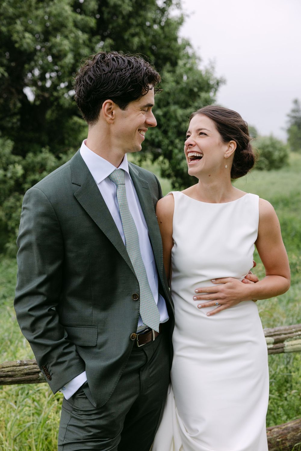 A couple in wedding attire share joyful laughter while standing outdoors in a natural setting.