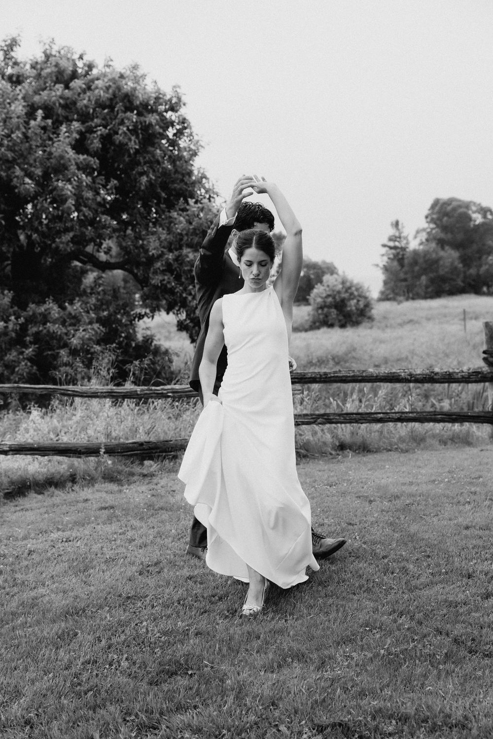 A bride in a sleek white dress twirls joyfully on a grassy field.