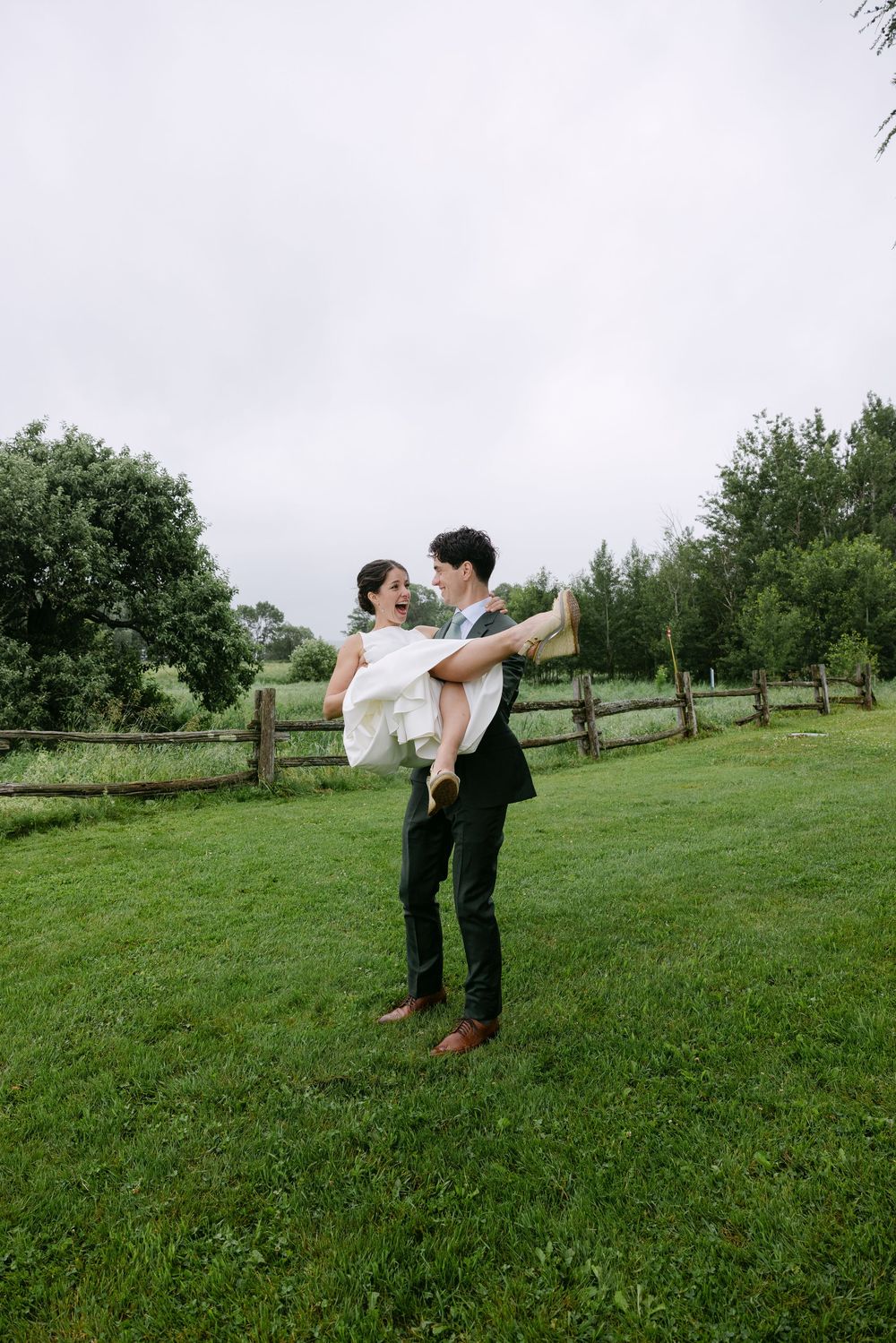 A groom lifts his bride playfully in the air on a green lawn surrounded by trees and fencing.