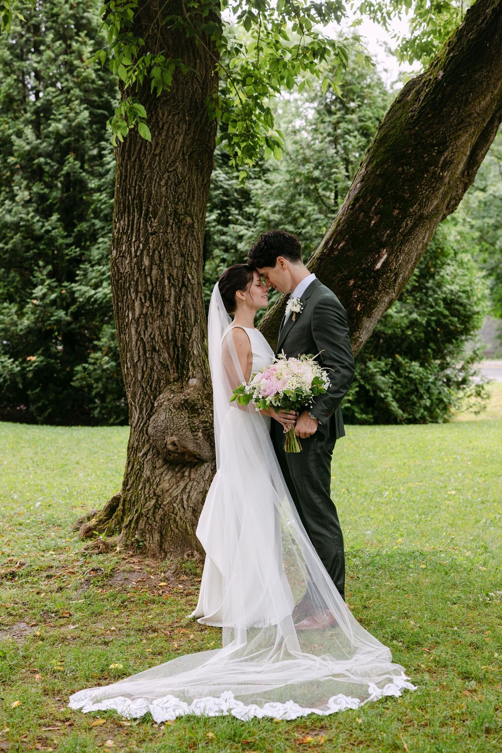 A couple in wedding attire share a romantic kiss under a large tree in a lush garden setting.